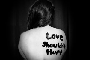 The bare back of a woman facing away from the camera. On her shoulderblade is written in black body paint the words "Love shouldn't hurt."
